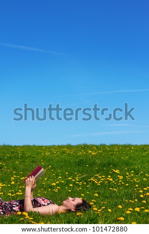young girl on a green meadow against blue sky reading a book