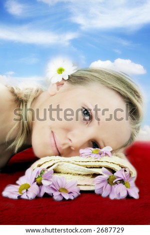a fresh beautiful blond woman laying on a red towel with flowers in front of the blue sky clouds