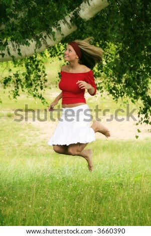 A young beautiful happy woman is jumping against a green tree and a meadow