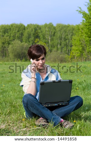 Girl sits on a green grass with a portable computer and talks by mobile phone