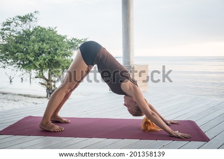 Young slim lady doing yoga downward facing dog on a beach with sunset over Bali in the background