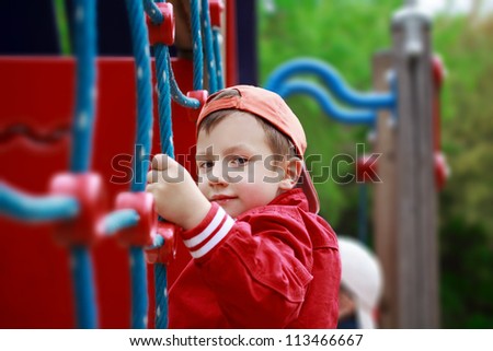 Cute boy in red jacket and cap climbing at jungle gym, brother in background blur