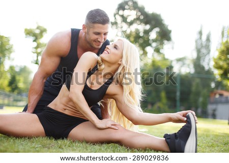 Young couple doing exercise outdoor on grass. Blonde woman stretching with personal fitness trainer