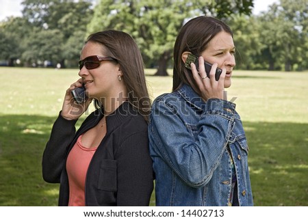two happy, beautiful young women talking on mobile phones in park, apparently unaware of each other