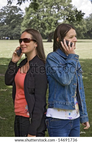 two happy, beautiful young women talking on mobile phones in park, apparently unaware of each other