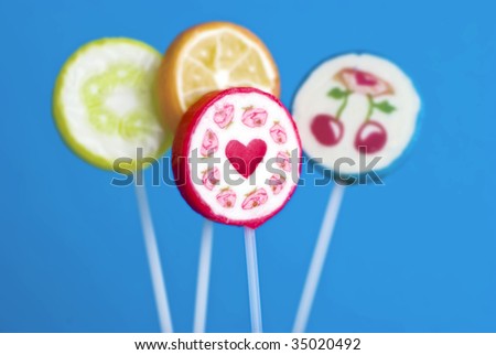 one sharp \'heart\' lollipop in front of three unsharp \'fruit\' lollipops; lollipops show heart surrounded by roses, lemon slices, orange slice and pair of cherries; all against blue background.