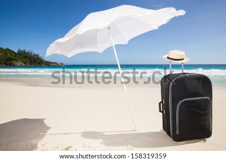 travel background with a black trolley suitcase  with a sun hat under a sunshade  at a tropical beach with a turquoise sea and a blue sky
