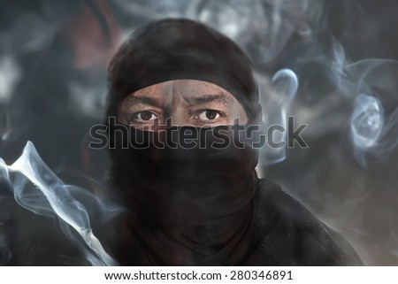 portrait of a black dressed and disguised male terrorist in a background of dark smoke. Concept for terror, terrorism, destruction and violence