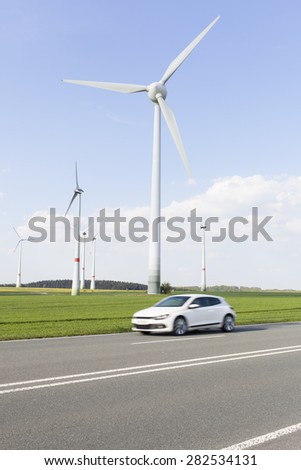 wind turbines near a country road with a fast white electric car passing by, concept for ecology, green energy, technology, progress, transportation and modern life