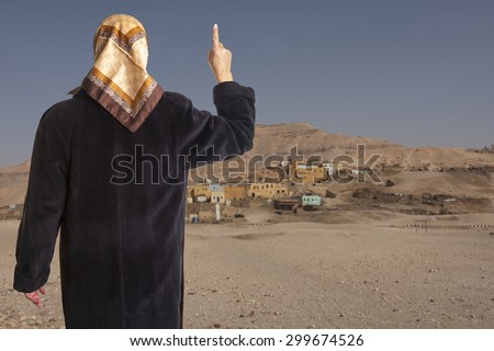 muslim woman dressed in a black coat and scarf showing the raised pointer finger of her right hand in front of an arabic village in the desert. Concept for islam religion and islamic state