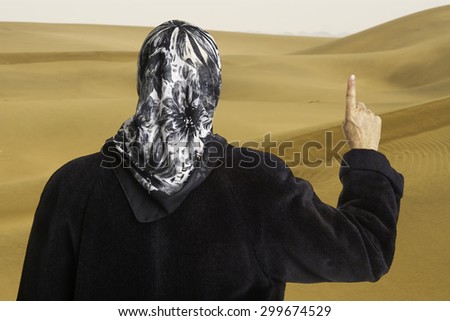 muslim woman dressed in a black coat and scarf showing the raised pointer finger of her right hand in front of sand dunes of Sahara desert. Concept for islam religion and belief
