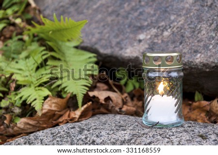 One lonely votive candle standing on tombstone and burning. Stone grave, green plants and old plant leaves seen in shallow background