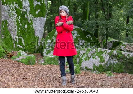 Young woman shivering with cold and embracing herself on a forest wearing a red overcoat, a beanie and gloves during winter