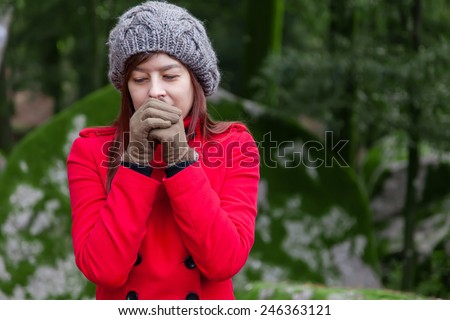 Young woman shivering with cold and blowing hot air to the hands on a forest wearing a red overcoat, a beanie and gloves during winter