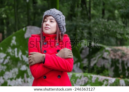 Young woman shivering with cold and embracing herself on a forest wearing a red overcoat, a beanie and gloves during winter