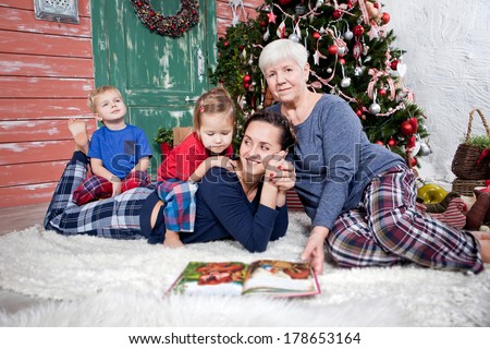 Happy family: grandmother, mother, little brother and sister sitting in pajamas at the Christmas tree with book for children
