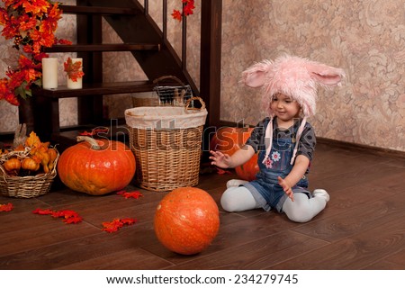 Cute baby girl in a funny hat with rabbit ears sitting on the floor next of different colorful autumn pumpkins. Halloween theme card, holidays