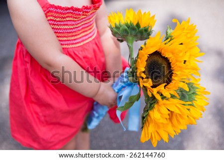 Unrecognizable toddler girl in a red dress with a bouquet of sunflowers in hands. Concept of style in children\'s fashion.