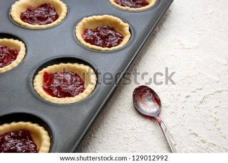 Filled, uncooked jam tarts in a tin, with a jam-covered teaspoon beside