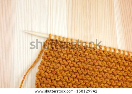 Garter stitch in deep orange yarn on a knitting needle, on pine board