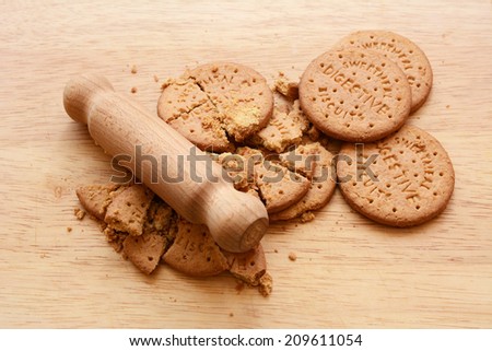 Whole and crushed digestive biscuits on a chopping board with a small wooden rolling pin