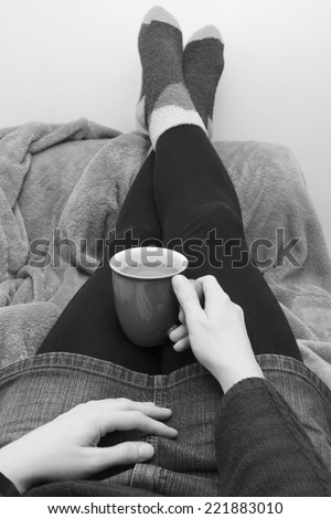 Woman dressed warmly, holding hot coffee or tea, with her feet up on the sofa - monochrome processing