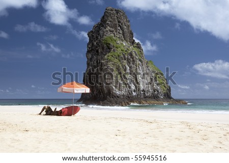 A surfer is laying under an umbrella waiting for waves. The deserted beach is shot against deep blue skies with few clouds and a huge rock in the middle. The picture is taken on Fernando de Noronha
