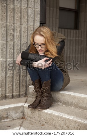 Outdoor photo of young teenage girl seated on ground, hugging knees, distraught facial expression.