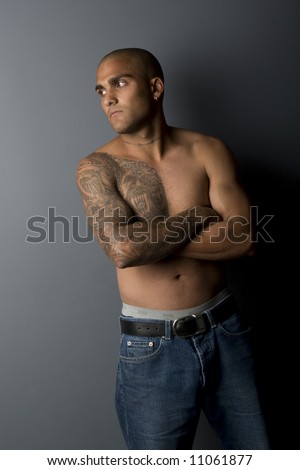 Shirtless attractive young ethnic man standing against gray wall looking sideways with arms crossed