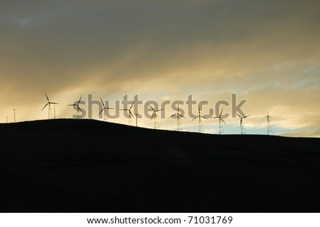 Row of silhouetted windmills on hillside with cloudy sky.