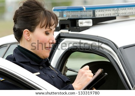 a female police officer writes a ticket while standing next to her patrol car.