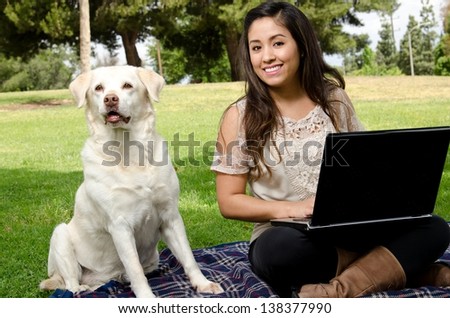 a smiling Hispanic woman sitting in the park with her dog and a computer.