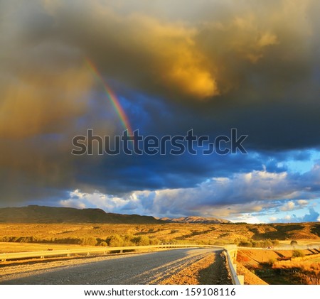 Low swirling cloud and flat plain covered in orange sunset. Cloud crosses the rainbow. In the steppe runs a gravel road. Storm over the Pampas