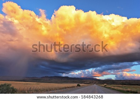 Summer rain. The huge cumulonimbus cloud is shined with the sunset sun. Rain streams shine orange light. The cloud hangs over the gravel road