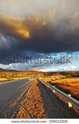 Low swirling cloud and flat plain covered in orange sunset. Cloud crosses the rainbow. In the steppe runs a gravel road. Storm over the Pampas