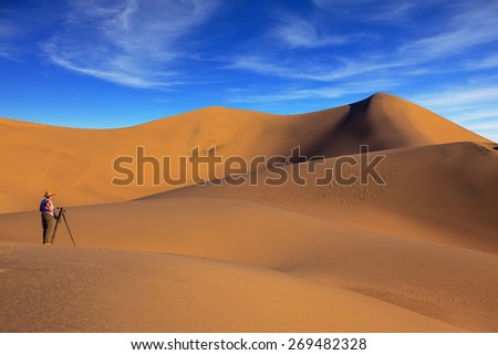 Sunrise in the orange sands of the desert Mesquite Flat, USA. Woman - photographer in a striped T-shirt is ready to shoot with a tripod among the sand dunes