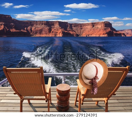 Waves from the boat cut through the Lake Powell. At the stern of the vessel are two deck chairs. On the back of one hanging elegant ladies straw hat.