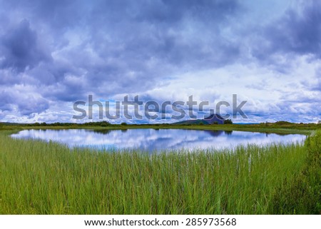 Small lake surrounded by green meadow. Summer Iceland. In the smooth water of cold lake reflects cloudy sky