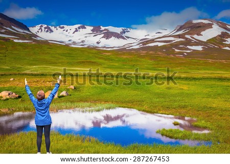 Blue lake water reflects the snowy hills. Iceland. The woman - tourist in blue jacket raised her hands in delight beauty of nature