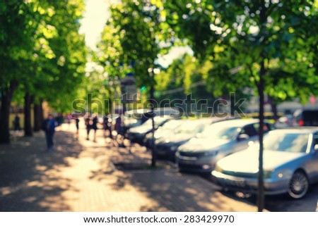 People walk through the alley between green trees in a park near the parked cars on a sunny day. Blurry