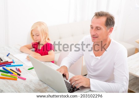 A father working on a laptop computer while his daughter is drawing.