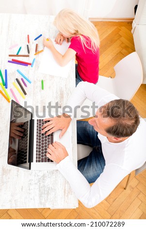 A father working on a laptop computer while his daughter is drawing.