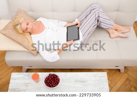 A mature woman with tablet enjoying cherries on the sofa seen from above.