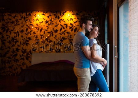 A young couple standing at home at the window while hugging in love.