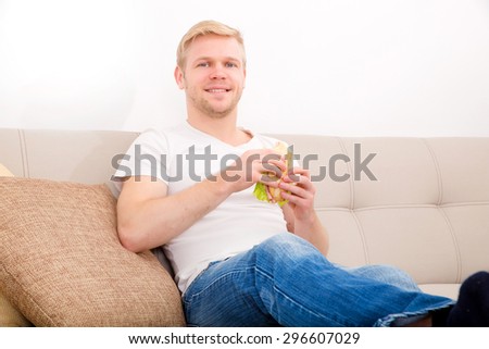 A young adult man eating a sandwich at home on the couch.
