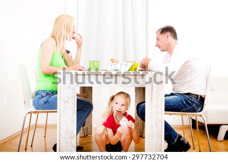 Girl eating chocolate beneath table while the family is having breakfast