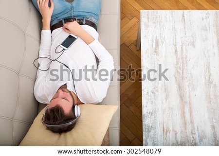 A young man lying on the sofa listening to music with some headphones and a smartphone.