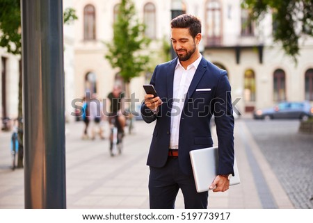 A handsome young businessman holding his laptop while using his phone and standing on the street