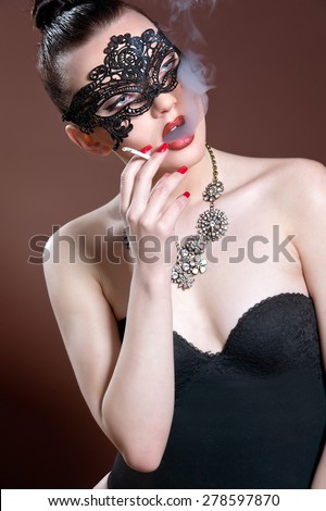 Fashion photo of gorgeous woman with dark hair and blue eyes, with lace mask on her face,posing in dark studio. Sexy Woman. Smoking Woman.