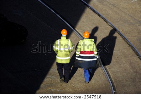 Two manual workers wearing colorful safety gear and helmets viewed from a high viewpoint walk away from camera from sunshine to shade